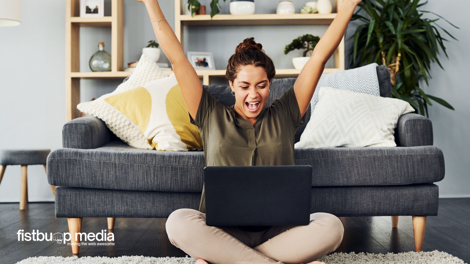 Woman sitting on the floor in front of a couch, arms raised in excitement while looking at a laptop screen
